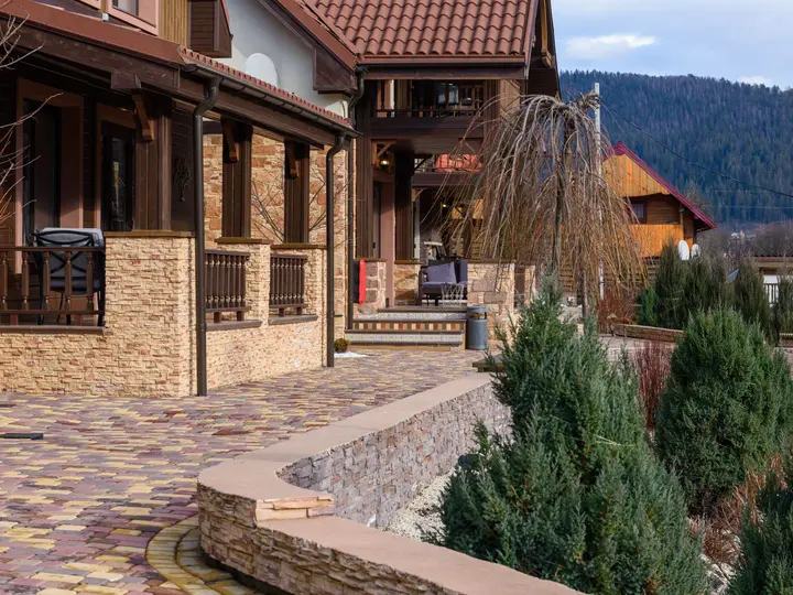 Landscaped front walkway of a stone and timber home, with a paver path, low stone wall and clipped evergreens