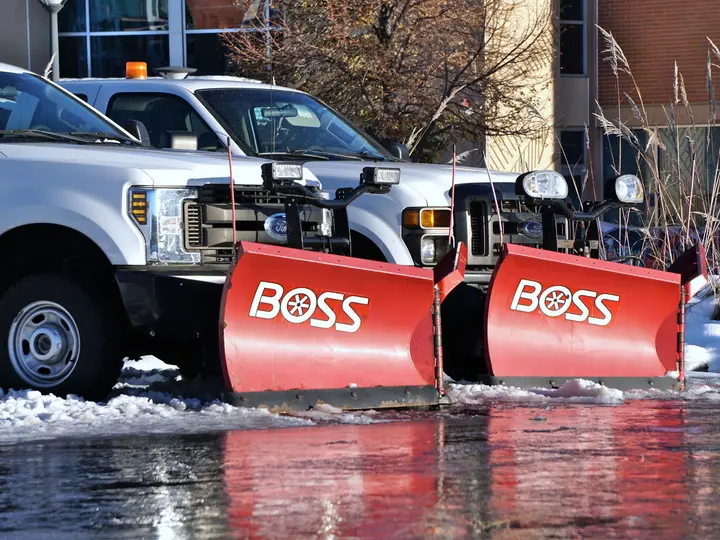 Two white pickup trucks fitted with red snow plow blades parked in a snow-covered lot