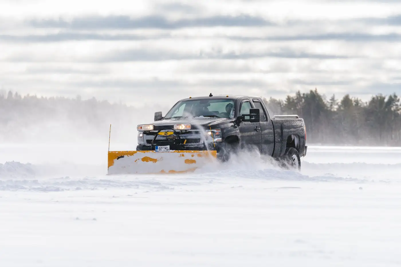 Pickup truck with a plow blade pushing a spray of snow across an open winter lot
