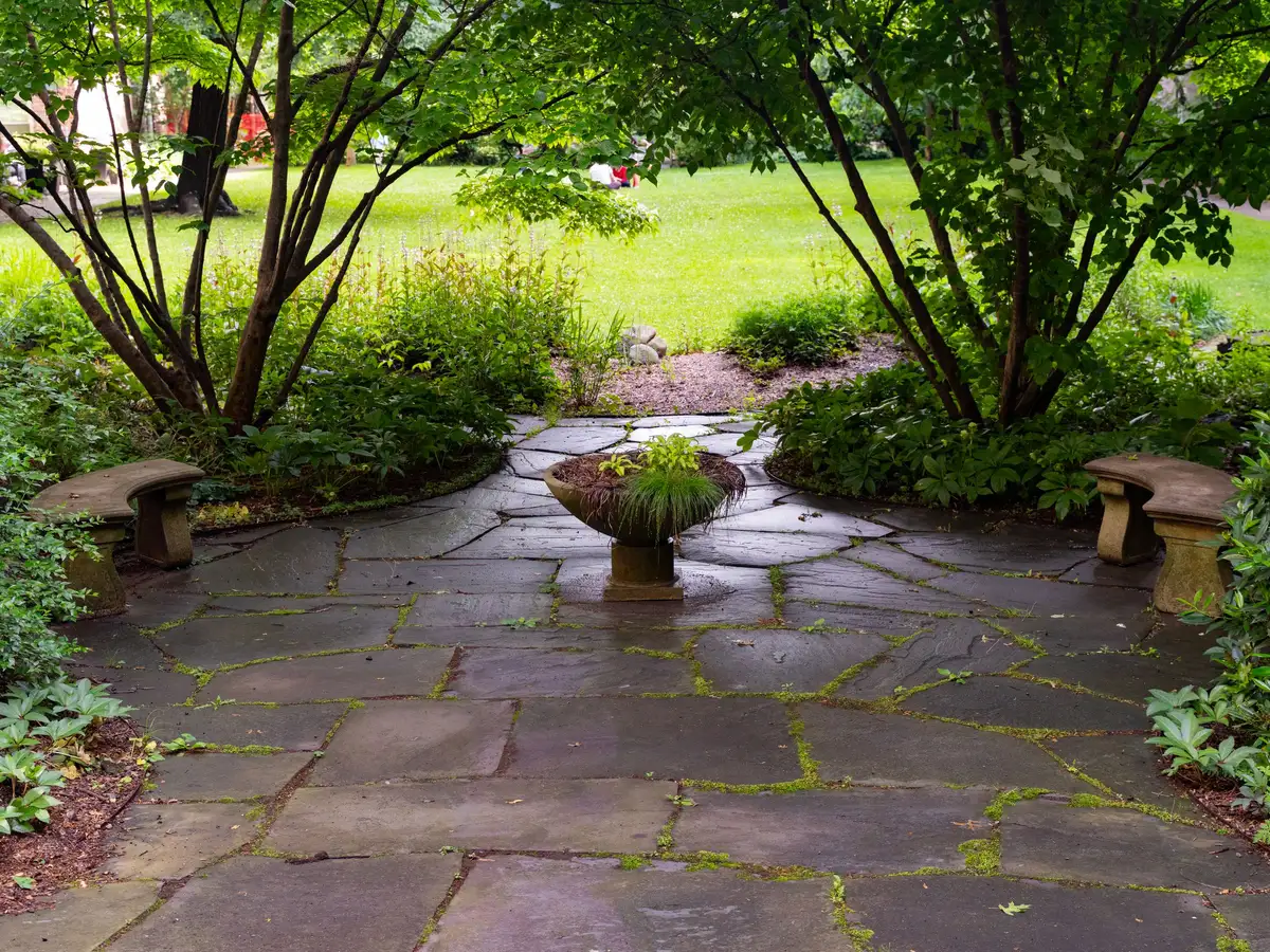 Natural stone patio and path with a stone pedestal, benches and dense garden plantings on either side